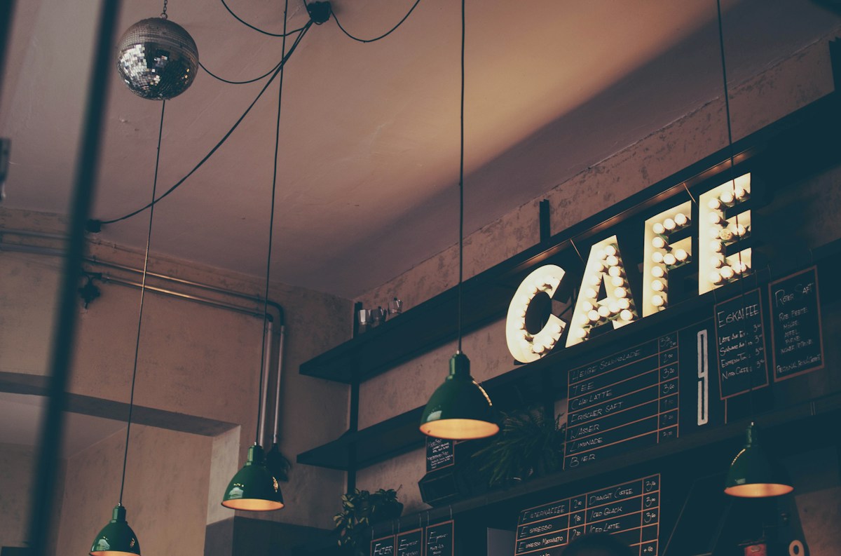 A warmly lit café sign above a chalkboard menu and green pendant lights